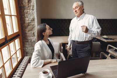 Two people in an office, one seated at a desk and another standing or sitting nearby, appearing to have a conversation